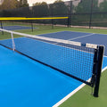 A close-up of a pickleball court featuring the Short Pickleball Net (31" H x 21'9" L) with crisp white lines on a blue and green surface; a chain-link fence and trees can be seen in the background.