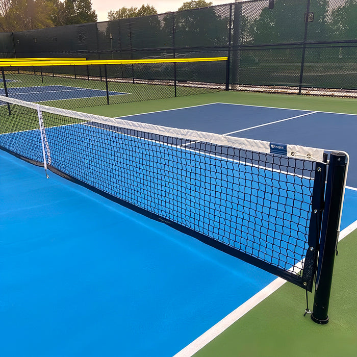 A close-up of a pickleball court featuring the Short Pickleball Net (31" H x 21'9" L) with crisp white lines on a blue and green surface; a chain-link fence and trees can be seen in the background.